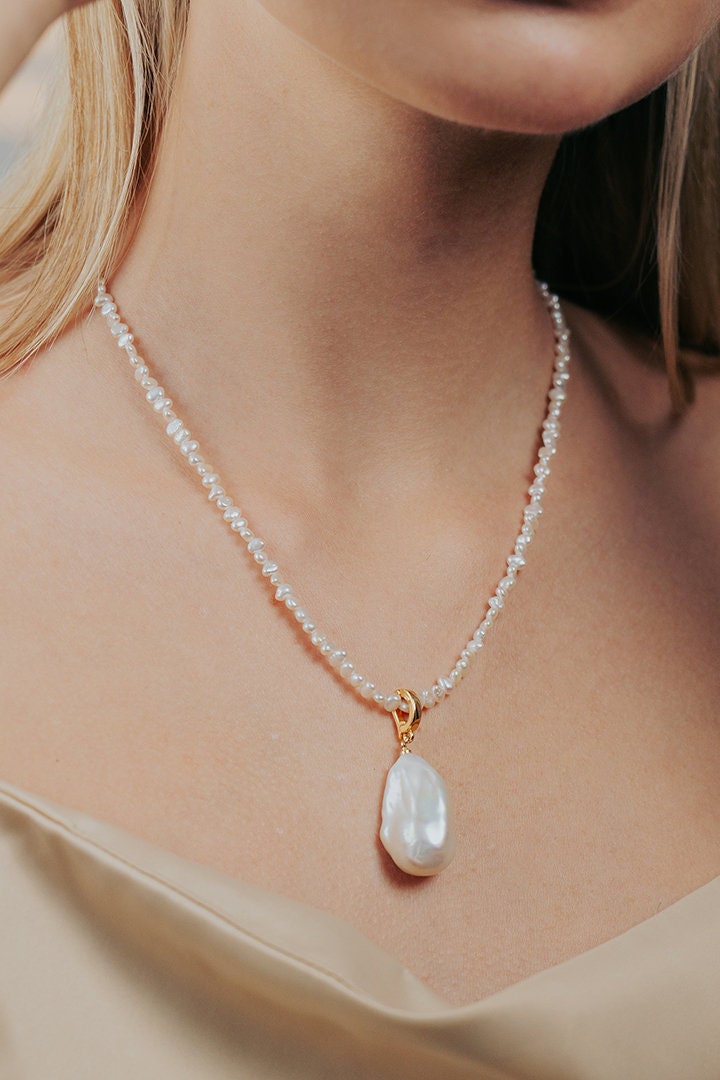 May include: A white pearl necklace with a large, irregular shaped, white pearl pendant. The necklace is made of small, white pearls and has a gold clasp.